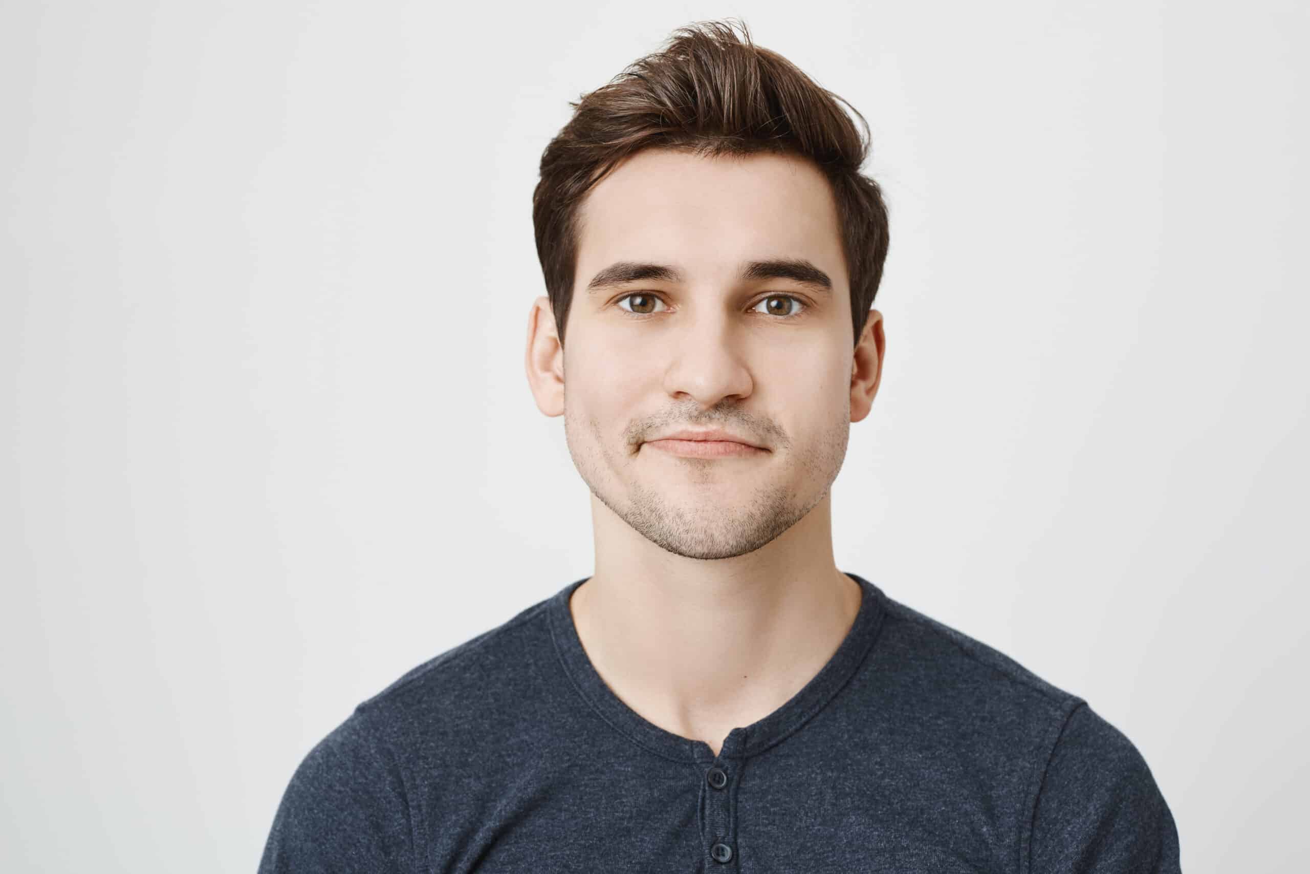 Close up portrait of nice european guy with stylish haircut and curvy smile, standing against gray background. Student came to his teacher to pass exam and feels awkward for not attending her classes